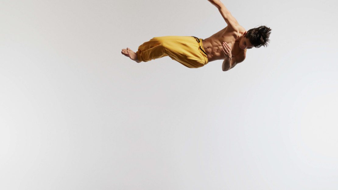 An athletic man performs a dynamic jump in a studio with a white background. The barefoot male wears yellow pants and displays defined abdominal muscles.