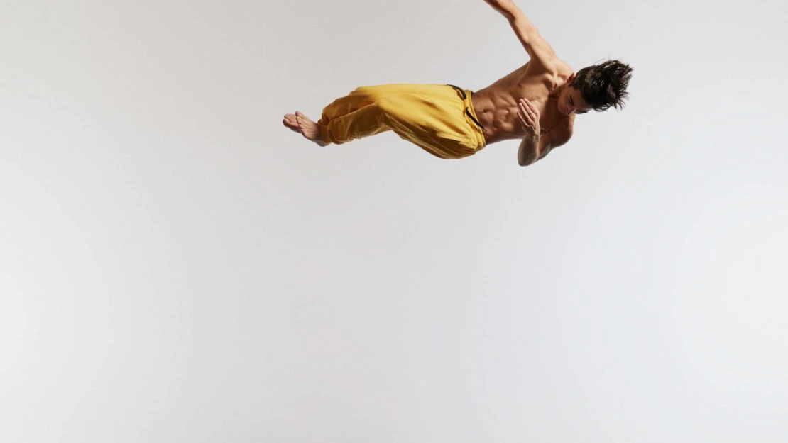 An athletic man performs a dynamic jump in a studio with a white background. The barefoot male wears yellow pants and displays defined abdominal muscles.