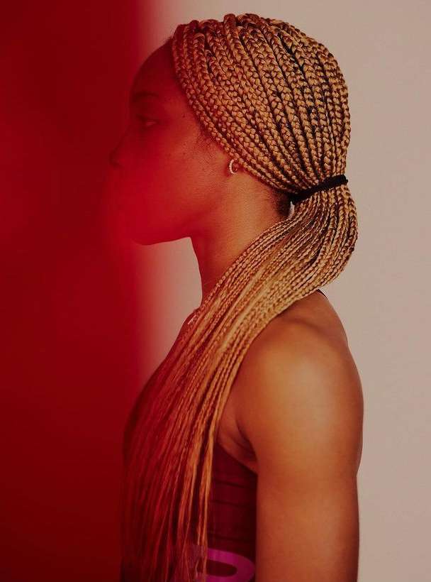 Profile of a tan-skinned woman with blonde braids, styled in a low ponytail, set against a split red and off-white background.