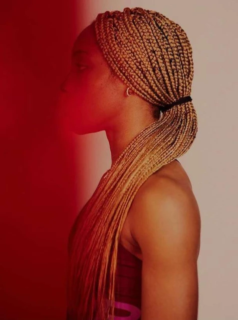 Profile of a tan-skinned woman with blonde braids, styled in a low ponytail, set against a split red and off-white background.