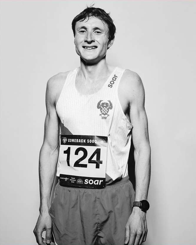 Black and white portrait of a young adult male runner wearing a race bib with the number 124, tank top, shorts, and a watch.