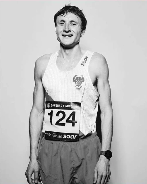 Black and white portrait of a young adult male runner wearing a race bib with the number 124, tank top, shorts, and a watch.