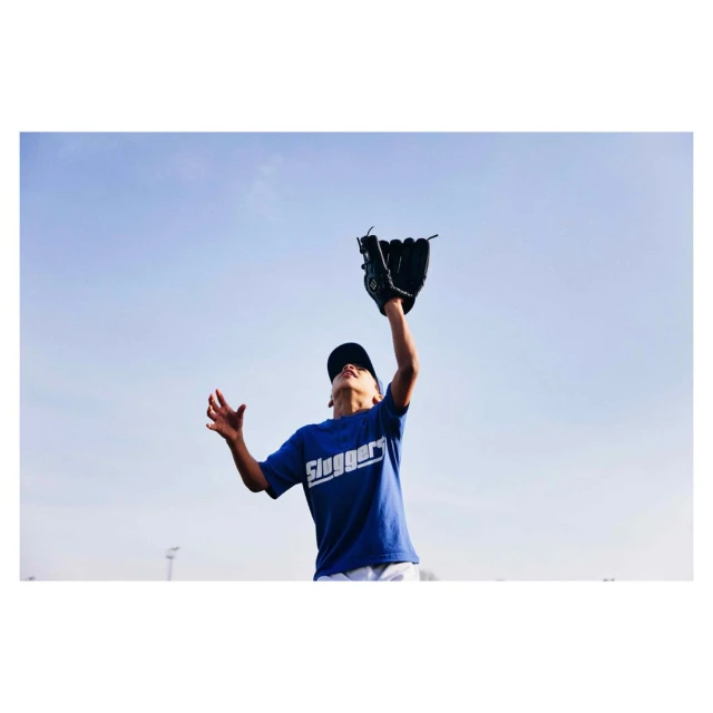 Young boy in blue Sluggers shirt reaches for a baseball with a glove against a clear blue sky in a bright, outdoor daytime shot.