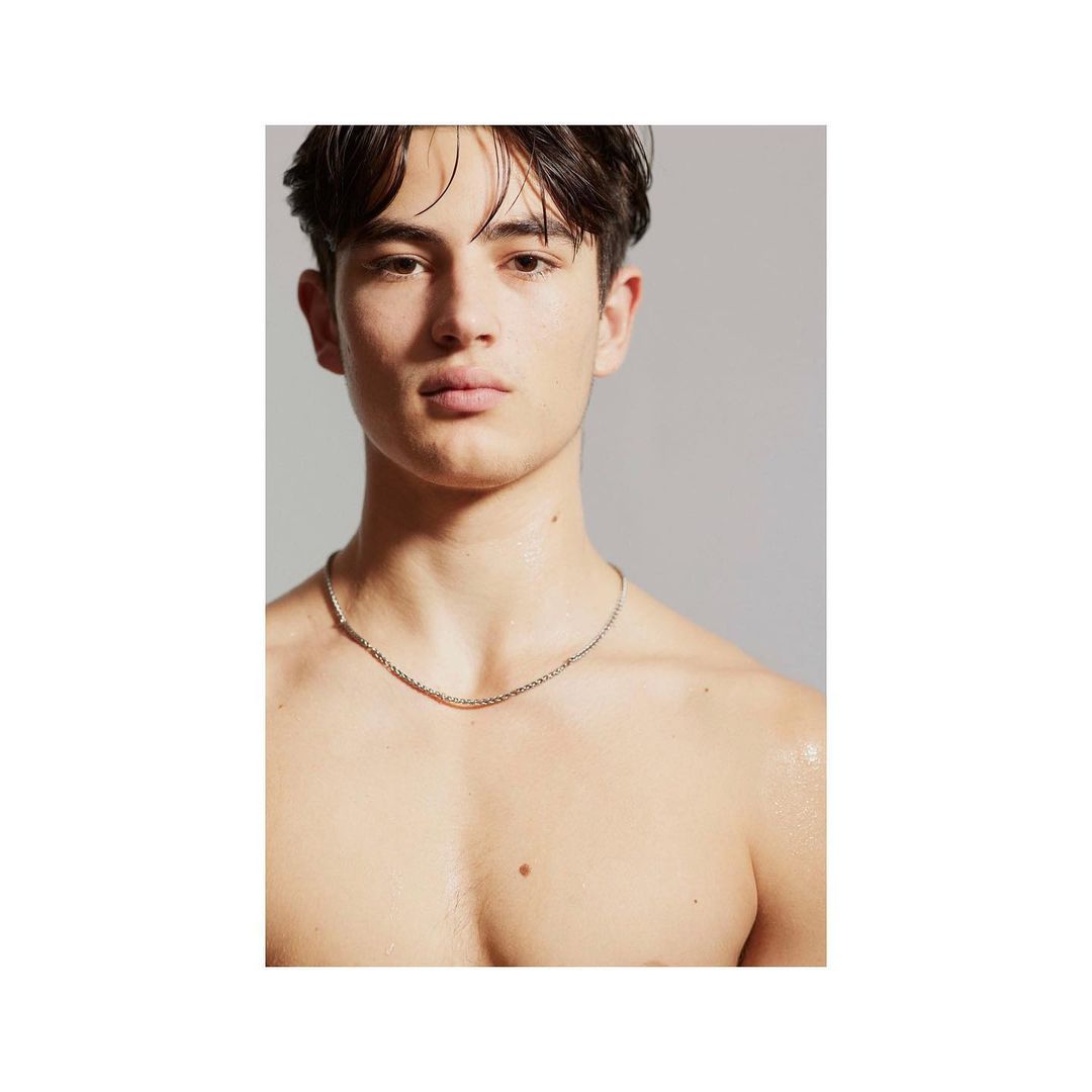 Close-up of a shirtless adult male with dark hair wearing a silver necklace in a studio with a grey background.