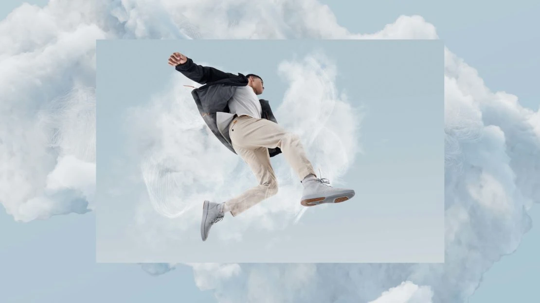 Adult male in mid-air wearing a jacket, t-shirt, pants, and sneakers in front of a cloud background in a fashion style image.