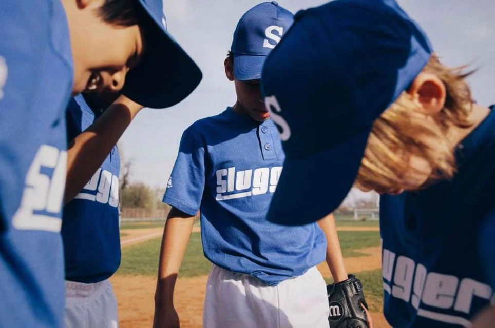 Group of young baseball players in blue uniforms with 'Slugger' on the front huddle together on a baseball field.