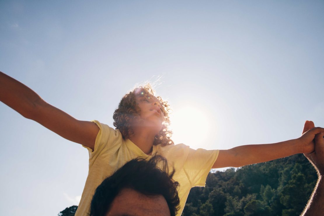 Child on adult's shoulders with arms outstretched against a sunny sky, framed by trees; freedom and summertime vibes in nature.