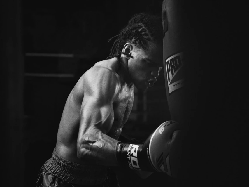 A muscular man with dreadlocks is shown in black and white, leaning against a punching bag in a boxing gym with boxing gloves on.