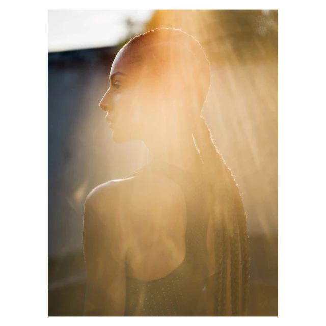Woman in profile bathed in golden sunlight with braids. Close-up portrait highlighting skin and hair texture against a blurred background.