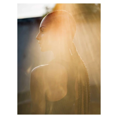Woman in profile bathed in golden sunlight with braids. Close-up portrait highlighting skin and hair texture against a blurred background.