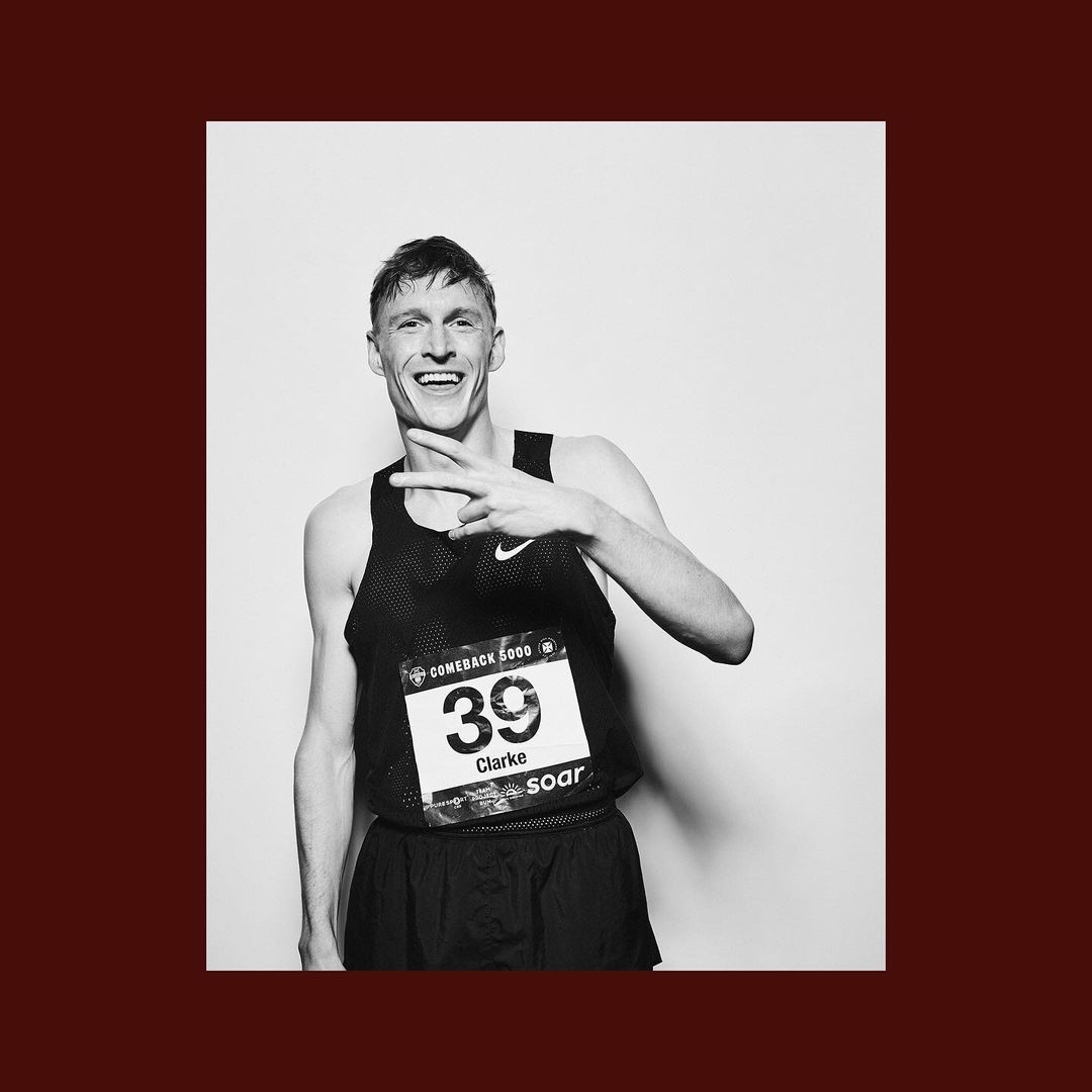 Black and white portrait of a smiling male athlete wearing a racing bib and tank top, making a peace sign in a light studio setting.