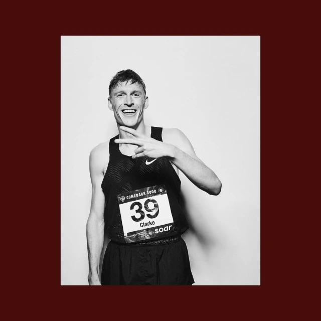 Black and white portrait of a smiling male athlete wearing a racing bib and tank top, making a peace sign in a light studio setting.
