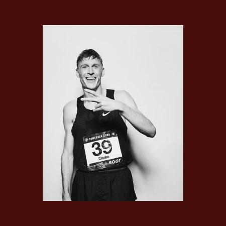 Black and white portrait of a smiling male athlete wearing a racing bib and tank top, making a peace sign in a light studio setting.