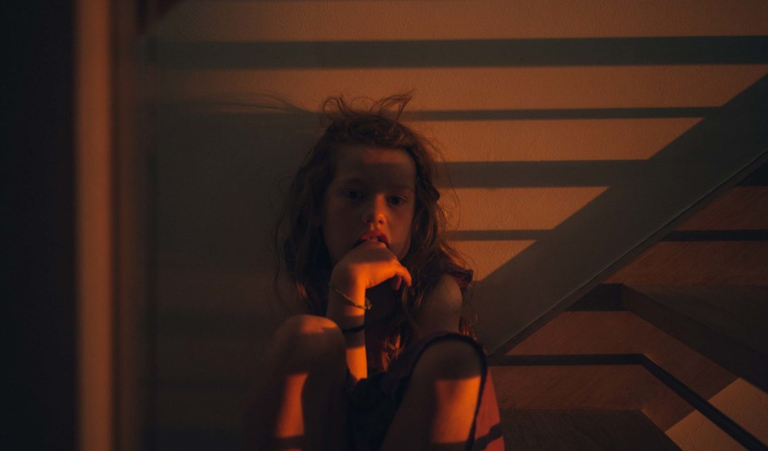 Child sits on stairs in the dark with orange lighting, wearing a bracelet, fingers to mouth, in an indoor environment.