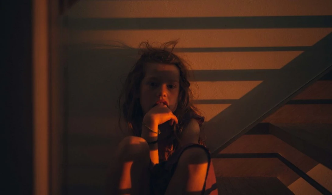 Child sits on stairs in the dark with orange lighting, wearing a bracelet, fingers to mouth, in an indoor environment.