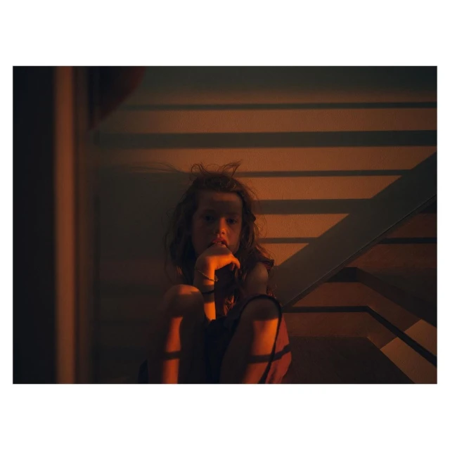 Young girl with disheveled hair sitting on stairs, lost in thought, in a warm-toned indoor setting with contrasting shadows.