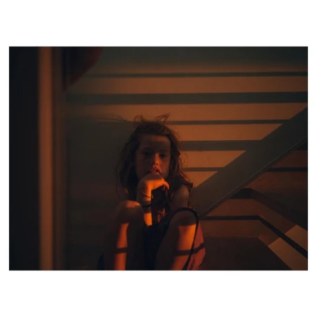 Young girl with disheveled hair sitting on stairs, lost in thought, in a warm-toned indoor setting with contrasting shadows.