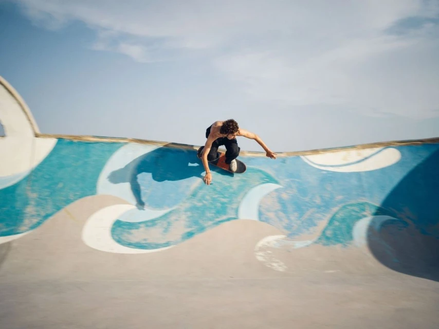 Young man skateboarding in a concrete skatepark bowl on a sunny day with a clear blue sky and abstract wave art in the background.