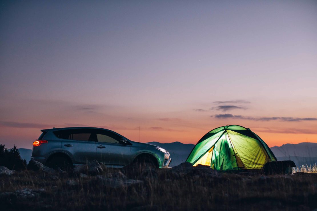 SUV parked near a lit tent at dusk, showcasing outdoor camping lifestyle under a twilight sky with distant mountains in the backdrop.
