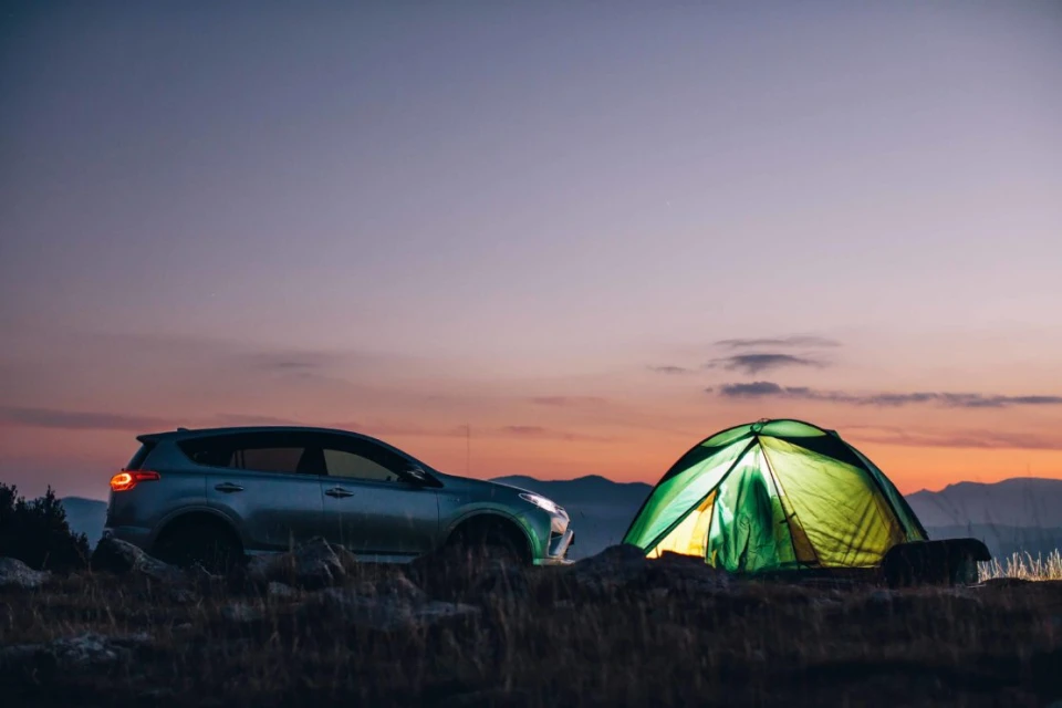 SUV parked near a lit tent at dusk, showcasing outdoor camping lifestyle under a twilight sky with distant mountains in the backdrop.