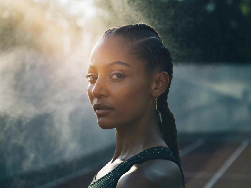 Close-up of a striking Black woman with cornrows, glancing at the camera with a neutral expression, athletic wear, sunlight, outdoor track.