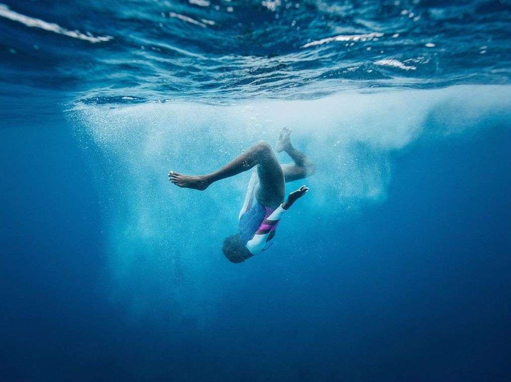 Underwater photo of a woman in a bathing suit, floating upside down in blue water with bubbles and sunlight.