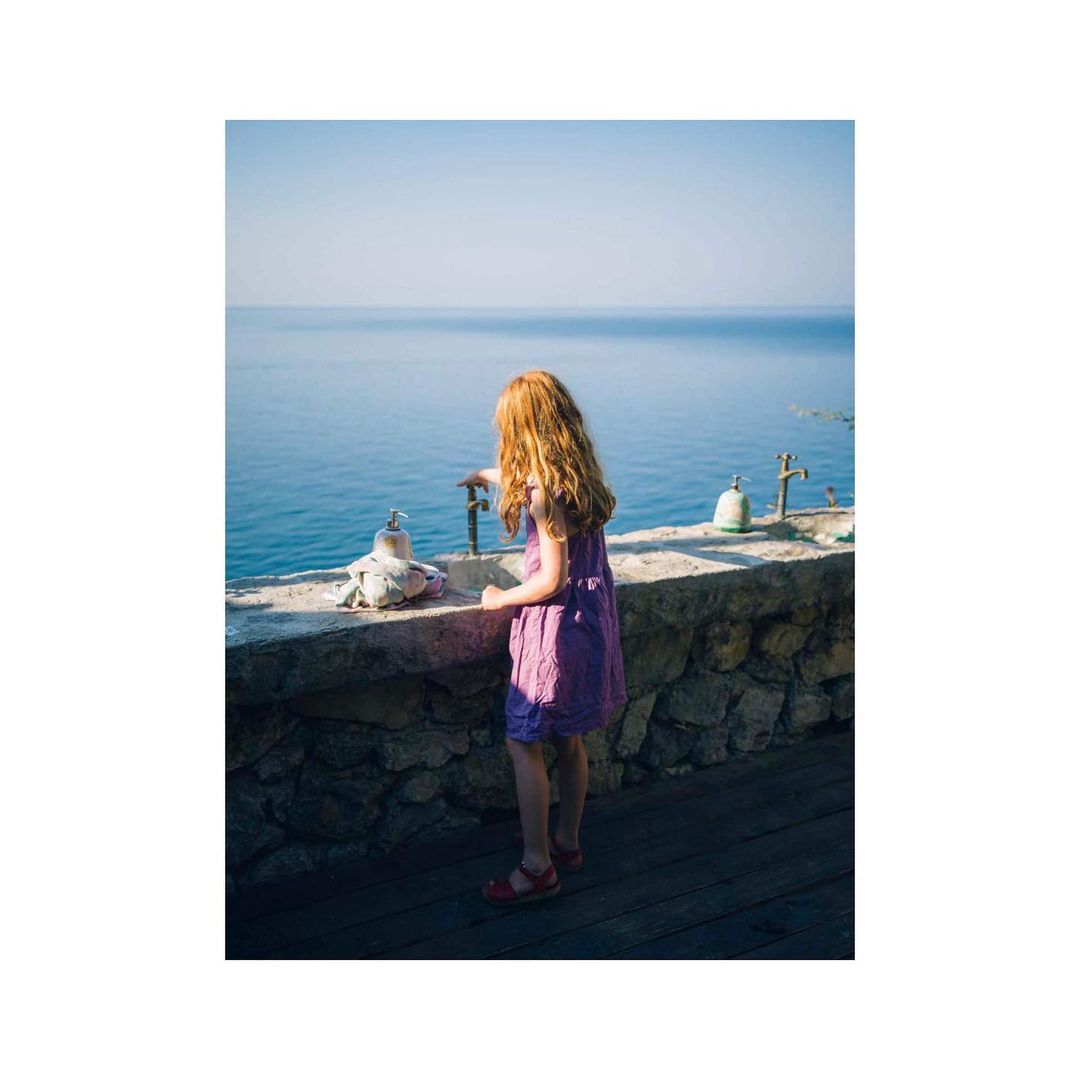 A young girl with red hair stands on a stone ledge overlooking a calm blue sea, wearing a purple dress and red sandals on a sunny day.