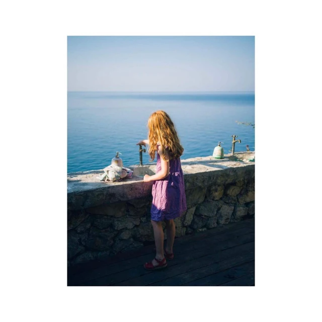 A young girl with red hair stands on a stone ledge overlooking a calm blue sea, wearing a purple dress and red sandals on a sunny day.