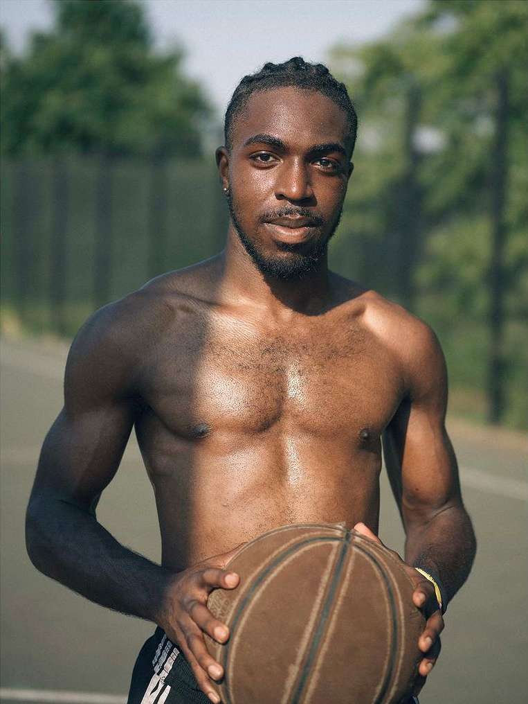 A shirtless athletic Black man holds a basketball in an outdoor setting, showcasing physical fitness and athletic lifestyle under natural light.