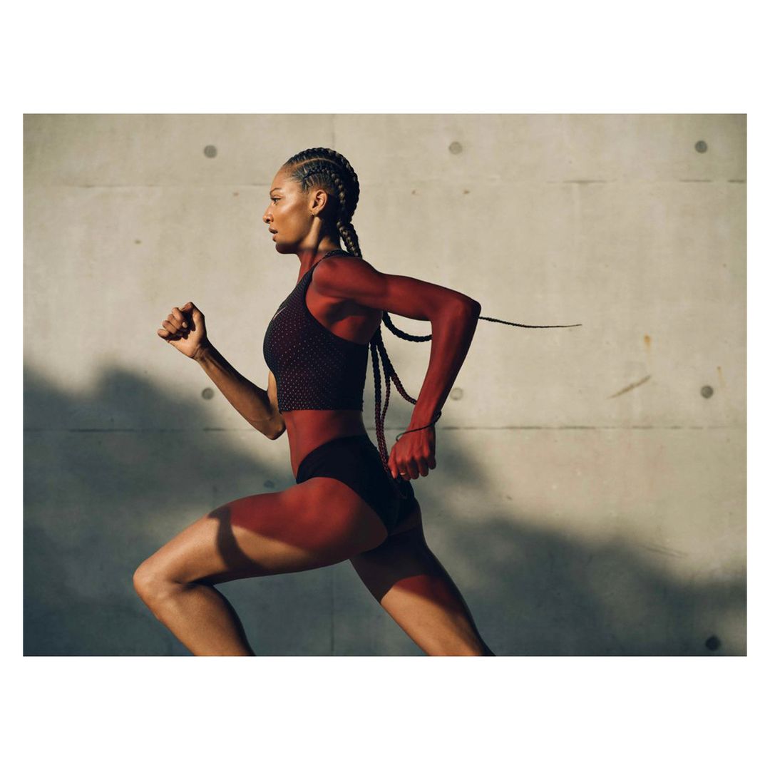 Athletic Black woman runs against a textured grey wall in a medium shot, wearing a sports bra and shorts, showcasing fitness and determination.