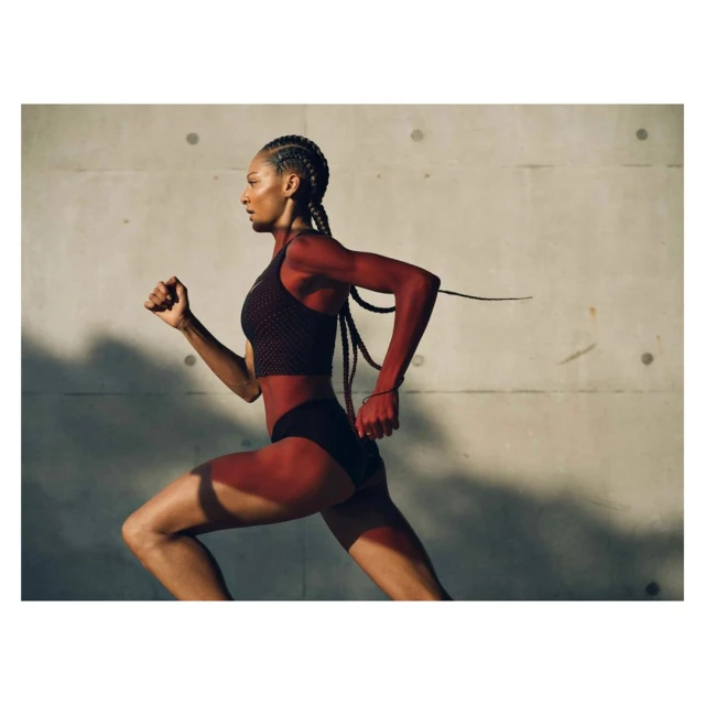 Athletic Black woman runs against a textured grey wall in a medium shot, wearing a sports bra and shorts, showcasing fitness and determination.