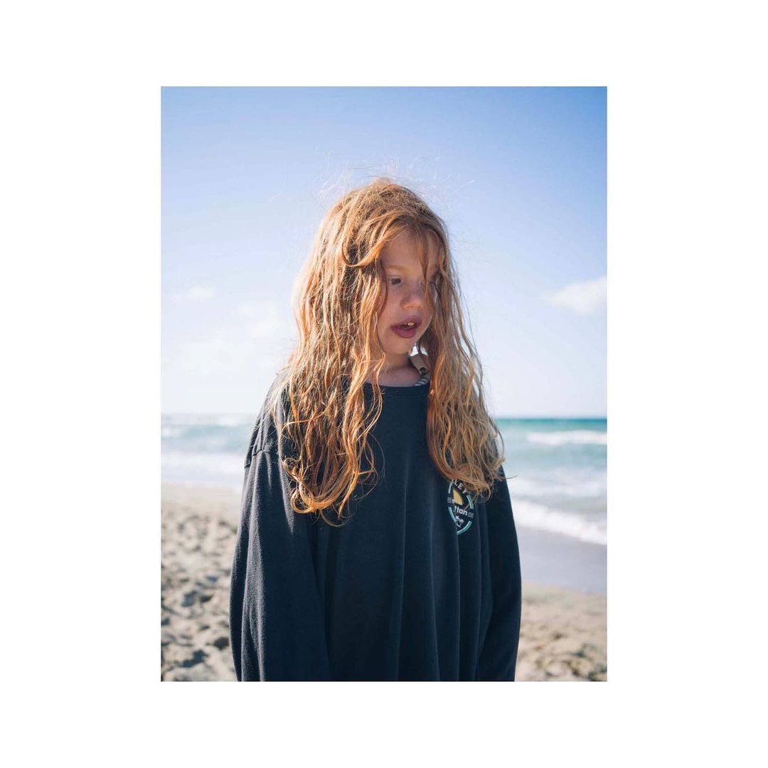 Young child with long curly reddish-brown hair wearing a dark long-sleeve shirt standing on a sandy beach near the ocean.