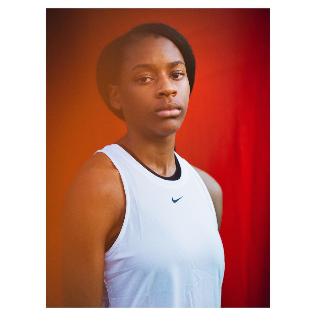 Portrait of a young African-American woman in a white tank top with a black Nike logo, posing against a vibrant red backdrop.