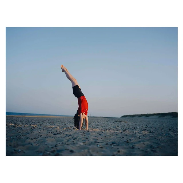 Young woman in red shirt and black shorts doing a handstand on a sandy beach with blue sky in background, showcasing fitness and athleticism.