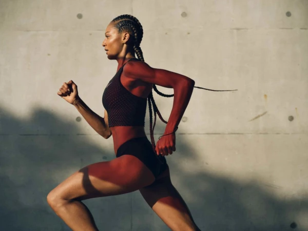 Black female athlete running in profile against a textured concrete wall, wearing a black sports crop top and briefs.