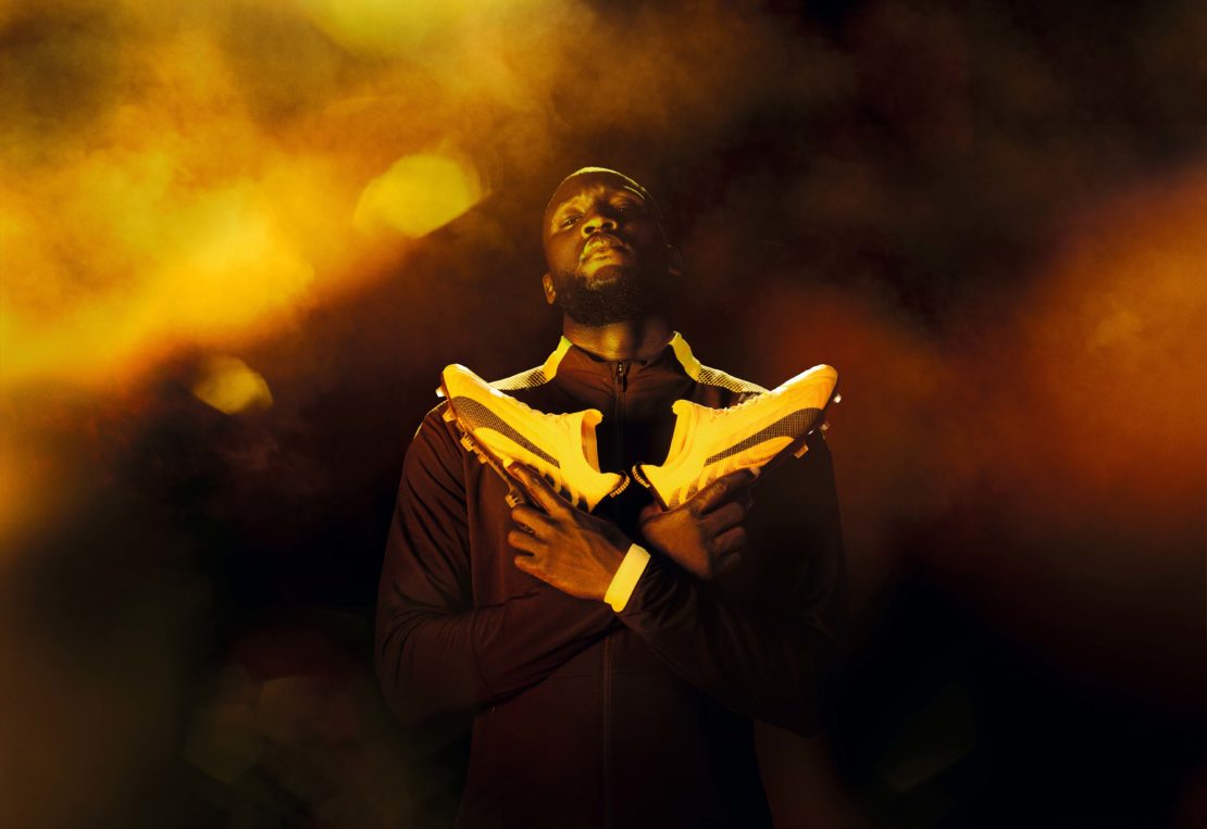 Adult male holding yellow and black soccer shoes in a studio with dark and golden background. Sport and fashion visual content.