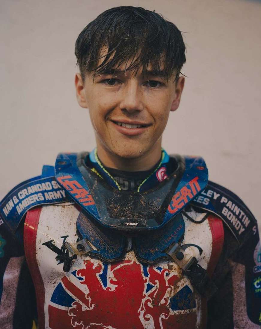 Portrait of a smiling young adult in motorsport gear, including neck brace with logos and patriotic flag design on the chest.