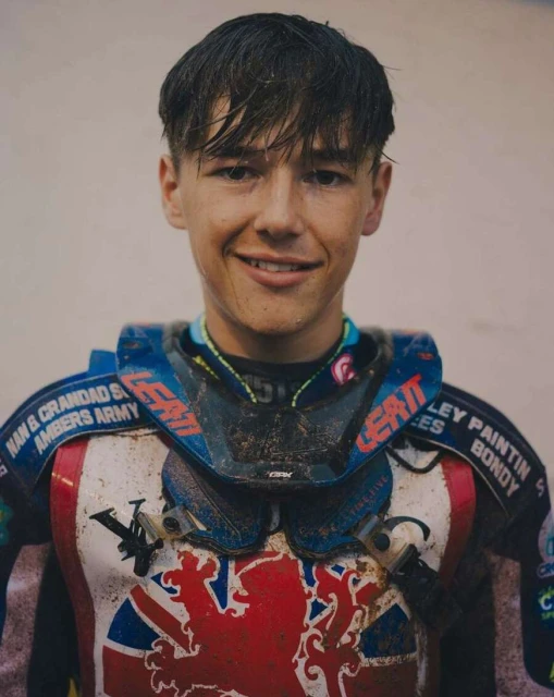 Portrait of a smiling young adult in motorsport gear, including neck brace with logos and patriotic flag design on the chest.