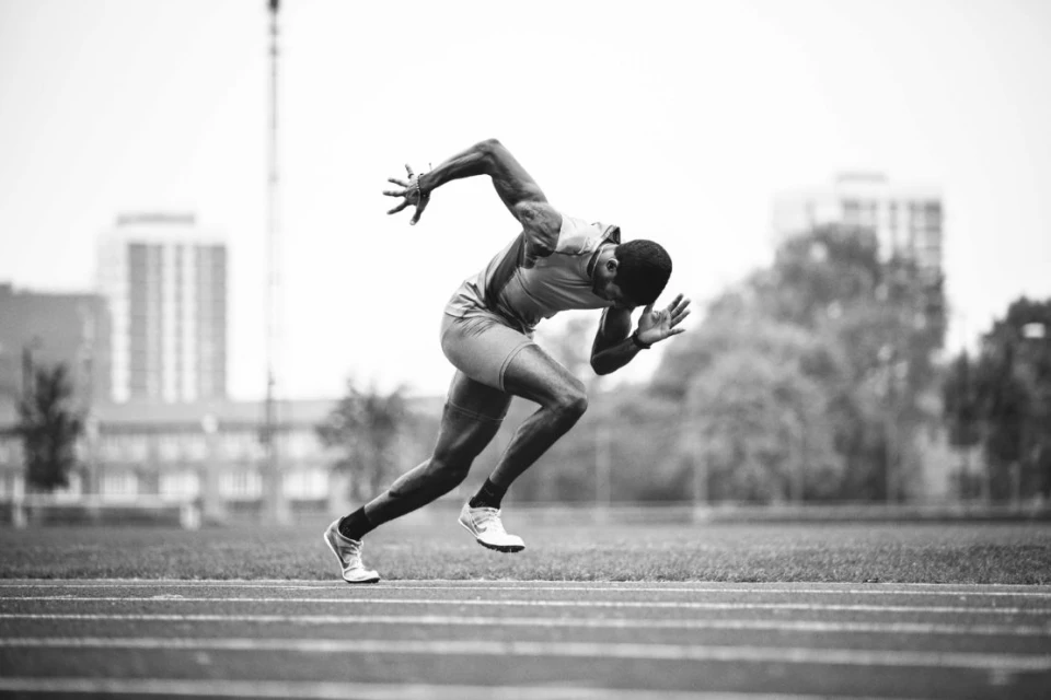Monochrome photograph of a muscular man in athletic wear sprinting on a track, showcasing determination and strength in an outdoor setting.