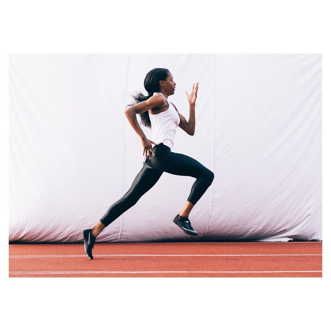 Athletic African-American woman sprints on an indoor running track with a white wall background wearing sportswear and running shoes.