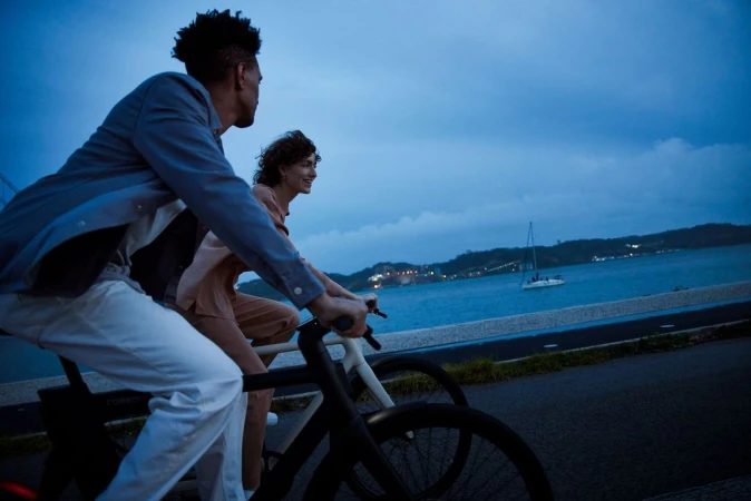 Man and woman riding electric bicycles along a coastal road at twilight, photographed by Paul Calver.