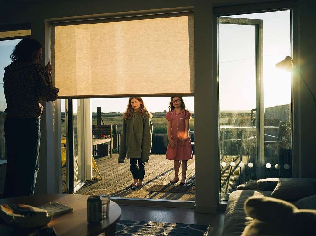 Indoor shot of a woman adjusting a window shade, revealing two young girls on a wooden deck near a grassy landscape on a sunny day.