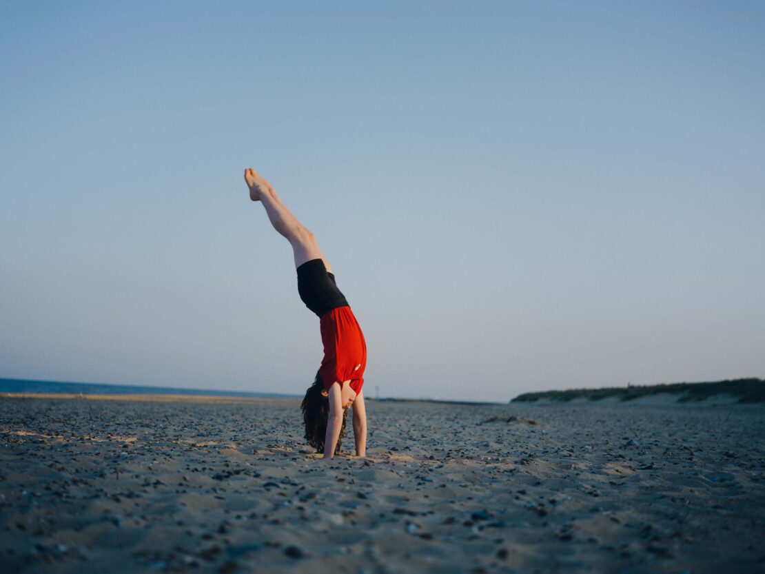 Young person performing a handstand on a sandy beach near the ocean under a clear sky. Athletic pose, outdoor fitness, sport.