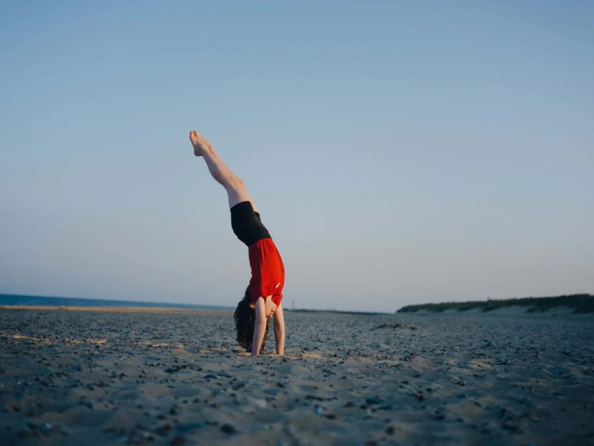Young person performing a handstand on a sandy beach near the ocean under a clear sky. Athletic pose, outdoor fitness, sport.