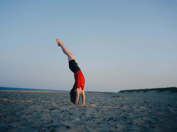 Young person performing a handstand on a sandy beach near the ocean under a clear sky. Athletic pose, outdoor fitness, sport.