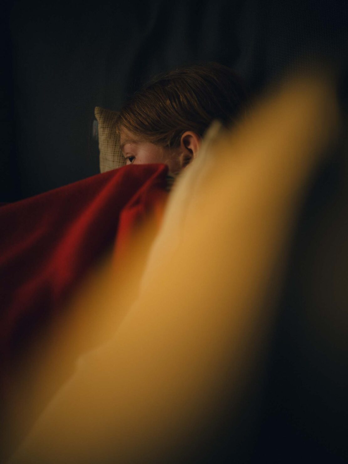Young child covered in red blanket, resting indoors. Soft lighting accentuates cozy indoor environment, hinting at a moment of relaxation.
