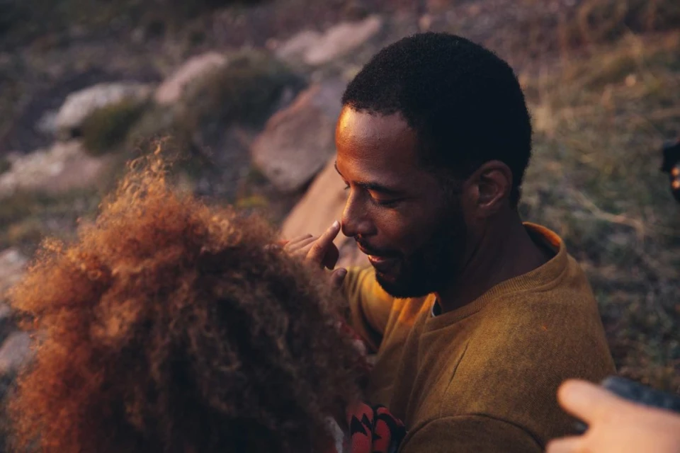 An African-American adult male touches noses with an auburn-haired adult female outdoors in a close, romantic moment, with a rocky backdrop.