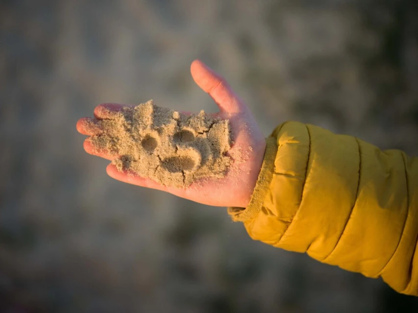 Child's hand holding sand with carved smiley faces in outdoor setting, focus on the sand and hand, with a blurred background.