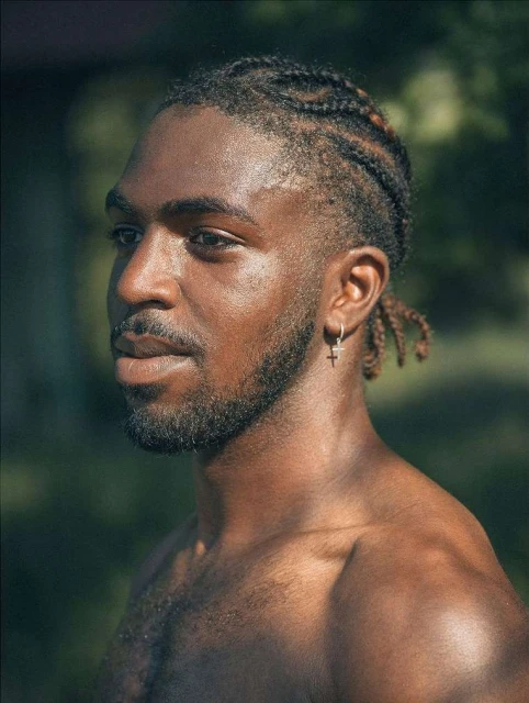 Portrait of an African American man outdoors. He has braided hair, light facial hair, and wears a silver cross earring.