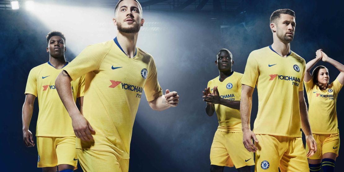 Group of six soccer players in yellow uniforms, standing against a dark blue background with bright overhead lighting. Team apparel.